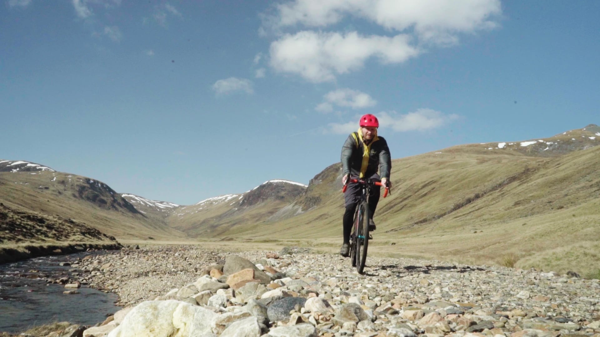 Stefan Morrocco riding a gravel bike along a stony riverside track in a Highland glen, with snow on the hills behind
